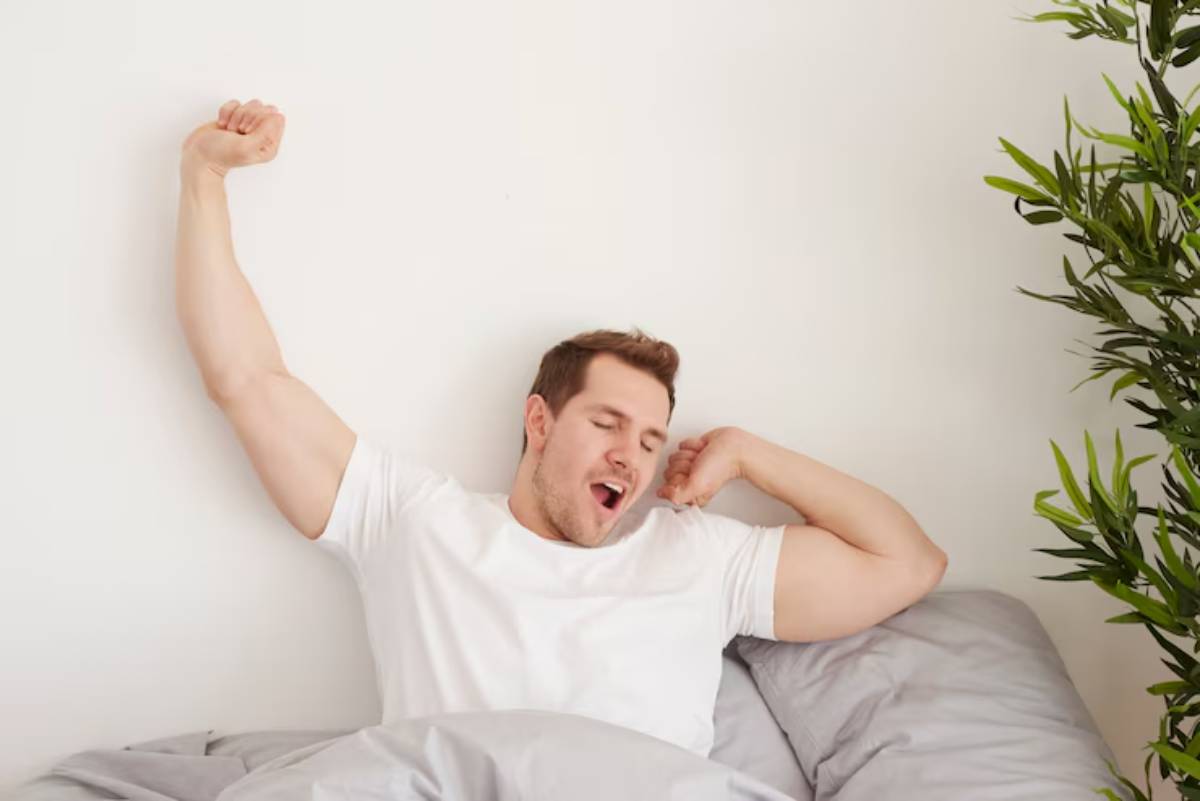 A person stretches in bed, wearing a white t-shirt, with gray blankets and a green plant in the background.